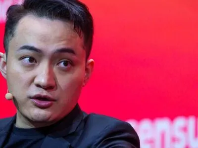 Джастин Сан в черном пиджаке и черной футболке, с маленьким бежевым микрофоном и наушником, на фоне красной стены