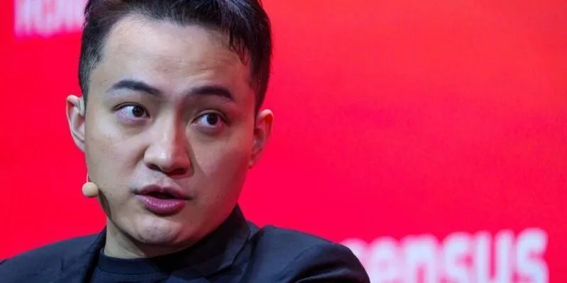 Джастин Сан в черном пиджаке и черной футболке, с маленьким бежевым микрофоном и наушником, на фоне красной стены