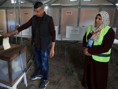 В городе Дейр-эль-Балах в центре сектора Газа, единственном городе, где организованы эти выборы, соорудили временные участки для голосования. Мужчина опускает бюллетень в урну, за этим наблюдает женщина в светоотражающем жилете и платке