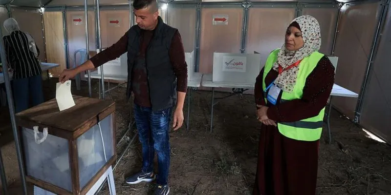 В городе Дейр-эль-Балах в центре сектора Газа, единственном городе, где организованы эти выборы, соорудили временные участки для голосования. Мужчина опускает бюллетень в урну, за этим наблюдает женщина в светоотражающем жилете и платке