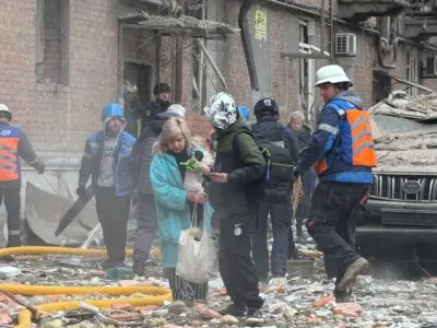 Женщина с собачкой в руках выходит из дома, на земле — обломки кирпичей, рядом с ней несколько человек в форме спасателей.