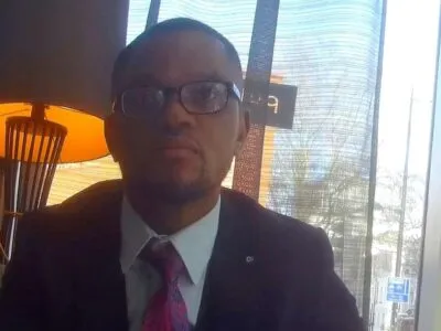 Фейковый юридический консультант, секретная съемка: темнокожий мужчина в очках, в пиджаке, белой рубашке и галстуке, на фоне окна и зажженной настольной лампы в лобби отеля.