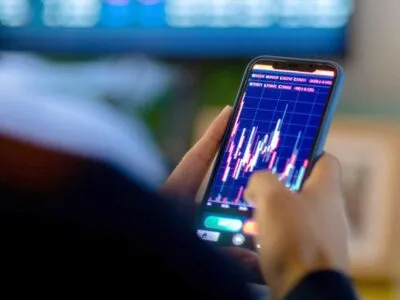 Man looking at trading app on his smart phone from his home office. He is being photographed from behind, so only his hands on his phone are visible in hard focus with the back of his shoulders and his office screens visible in soft focus.