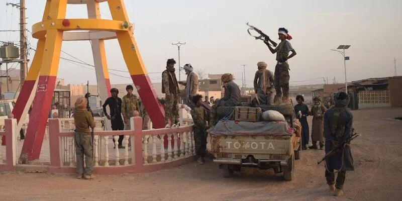 Группа боевиков в камуфляжной форме стоит вокруг пикапа на пыльной дороге. Одним из них стоит на автомобиле и держит в руках автомат. Слева на снимке видны коричнево-желтые ноги скульптуры.