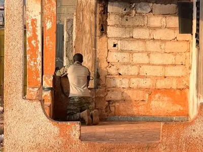 Мужчина в камуфляже с оружием стоит на коленях в кирпичном здании и наблюдает в окно