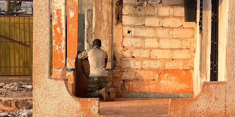 Мужчина в камуфляже с оружием стоит на коленях в кирпичном здании и наблюдает в окно