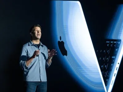 Джон Тернус на черном фоне, за спиной большое изображение макбука