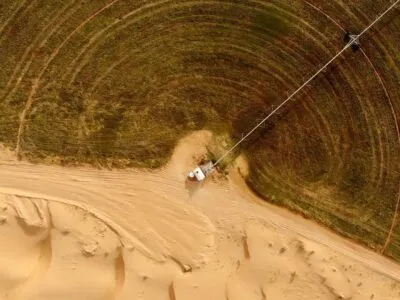 Вид с высоты на круглое зеленое сельскохозяйственное поле, граничащее с пустынным песком, с длинным трубопроводом, идущим от небольшого белого сооружения к зеленой зоне.