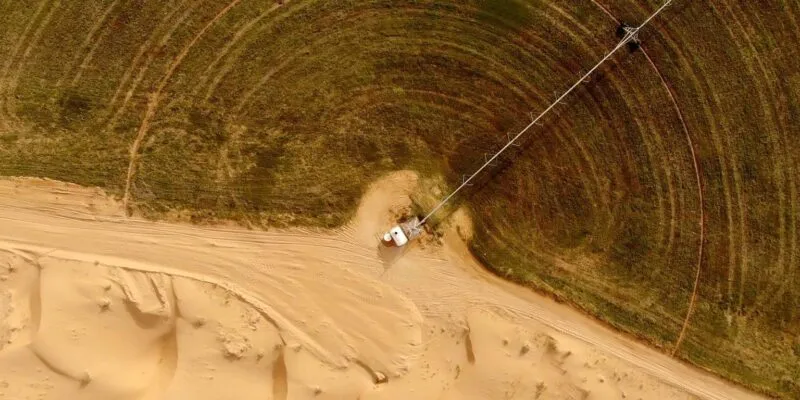 Вид с высоты на круглое зеленое сельскохозяйственное поле, граничащее с пустынным песком, с длинным трубопроводом, идущим от небольшого белого сооружения к зеленой зоне.
