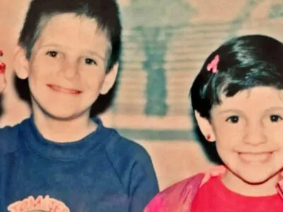 A boy in a blue top and a girl in a pink top, both with dark hair, smile at the camera - the boy is holding up a toy figurine