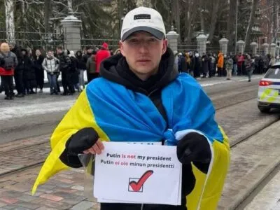 Роман Голиков стоит на улице с флагом Украины на плечах и плакатом с надписью «Putin is not my president»