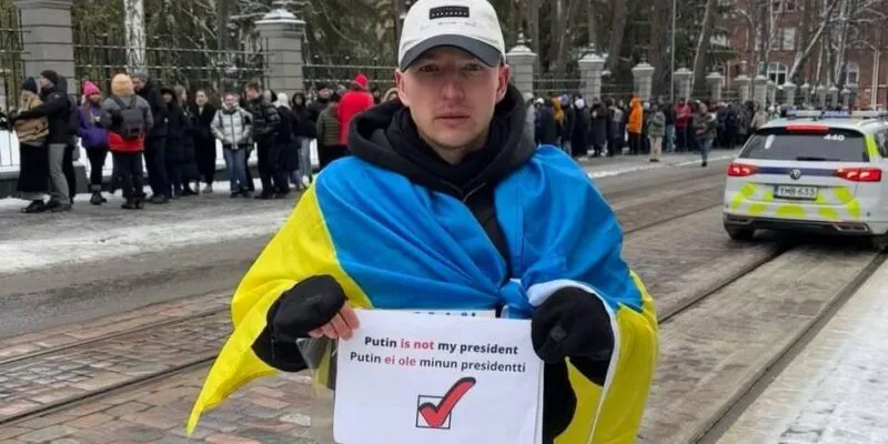 Роман Голиков стоит на улице с флагом Украины на плечах и плакатом с надписью «Putin is not my president»