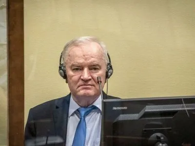 Ратко Младич в костюме с галстуком сидит за стеклом, на нем наушники, перед ним микрофон и монитор.
