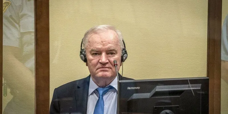 Ратко Младич в костюме с галстуком сидит за стеклом, на нем наушники, перед ним микрофон и монитор.