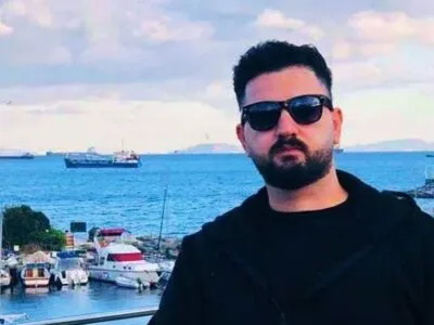 Kardo Ranya stands in the foreground on a waterfront balcony, wearing dark sunglasses, a black jacket, and a dark shirt. Behind him is a coastal marina with several small boats and yachts moored along docks, their white hulls contrasting with the deep blue water. Farther out on the sea, a few large cargo ships are visible near the horizon. To the right, a pier or harbour structure extends into the water, and low buildings line parts of the shore.