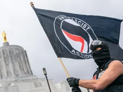 Мужчина в черной балаклаве держит в руках флаг антифа
