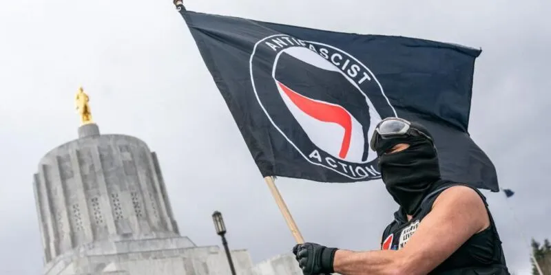 Суд в Техасе приговорил восемь активистов «Антифа» к крупным срокам за нападение на иммиграционный центр. Лидер группы получил 100 лет тюрьмы 1 Мужчина в черной балаклаве держит в руках флаг антифа