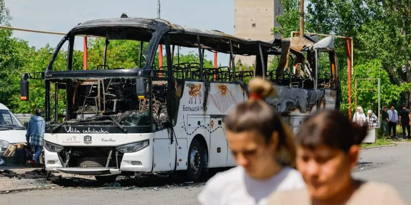 В самопровозглашенной ДНР заявили о восьми погибших при ударе по рейсовому автобусу. Что известно 1 Две женщины идут по улице, за ними — сгоревший автобус.