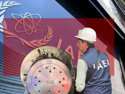 Человек в белой защитной каске и синем жилете с белыми буквами IAEA («МАГАТЭ») на спине. Он стоит рядом с металлическим цилиндром.