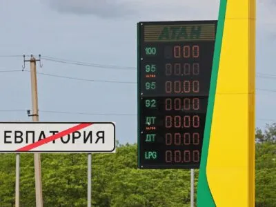 На заднем фоне темно-синее небо, зеленые деревья и столбы с проводами. На переднем плане слева дорожный знак о выезде из Евпатории и погасшее табло автозаправки "Атан".