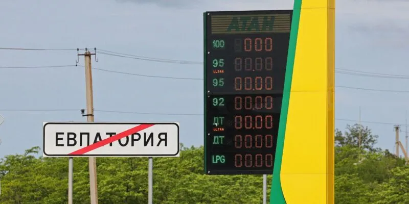 Локальный дефицит бензина. Как атаки на нефтяную инфраструктуру России повлияли на рынок топлива 1 На заднем фоне темно-синее небо, зеленые деревья и столбы с проводами. На переднем плане слева дорожный знак о выезде из Евпатории и погасшее табло автозаправки "Атан".