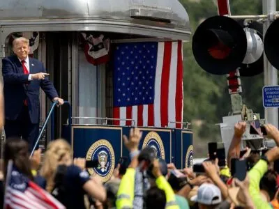 Трамп стоит в хвосте поезда с американским флагом, перед ним — много людей.
