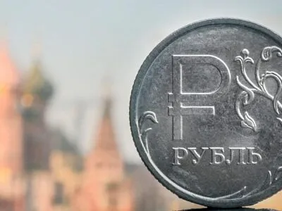 На заднем плане размытые очертания православного собора Василия Блаженного в Москве. На переднем плане монета из серого металла с надписью "рубль"