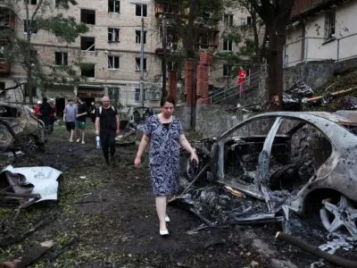 Люди идут между сгоревших машин, за их спинами поврежденный жилой дом