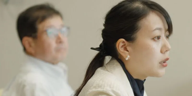 Сёко Кавата, мэр в белом блейзере и темной блузке, сидит в кресле. Позади нее виден ее заместитель в светлой рубашке