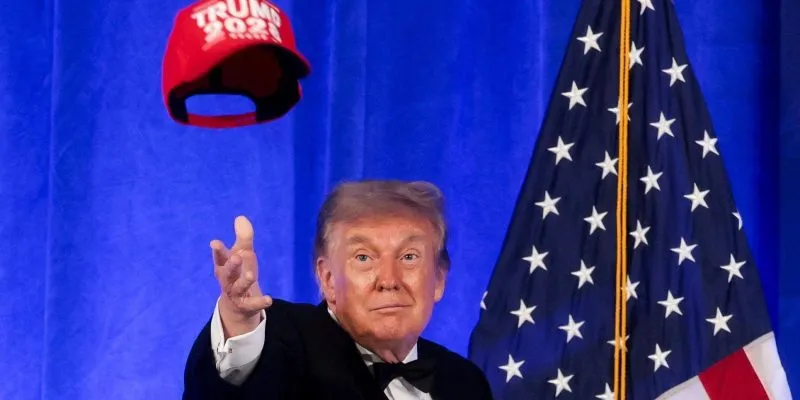 Трамп, кидающий в воздух красную кепку с надписью Trump 2028, за ним — флаг США.