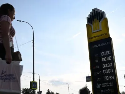 На заднем фоне — голубое небо. На среднем плане деревья, фонарные столбы и заправка «Роснефти». На табло АЗС нули напротив всех видов топлива, кроме дизельного. На переднем плане слева направо идет женщина с пакетом руке.