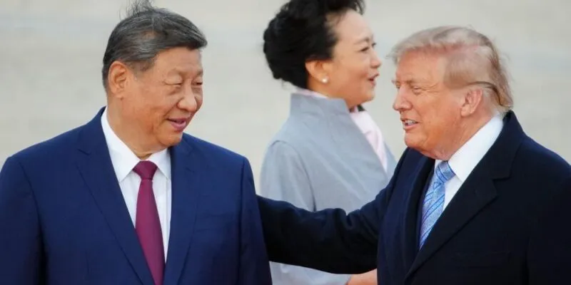 President Xi smiles and looks toward Trump, who has his hand on Xi's shoulder. Both are in suits and their haor is slightly tussled by wind. Xi's wife, in a light blue coat, looks away in the background.