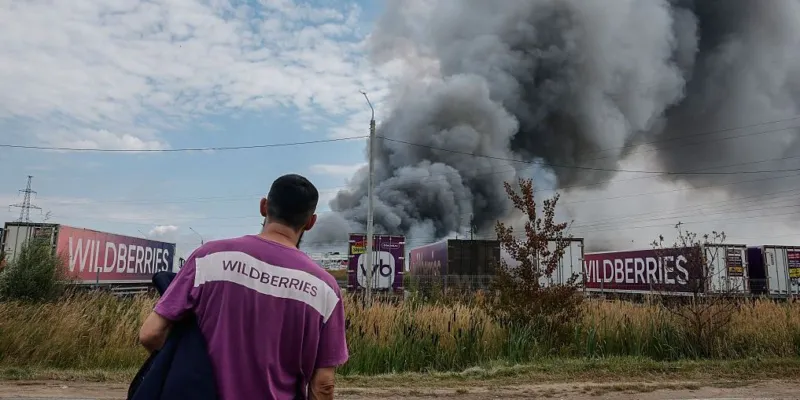 Человек в униформе маркетплейса с надписью Wildberries на спине наблюдает за клубами дыма над горящим складом