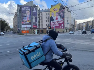 Правящие реформисты, неизбежность коалиции и «русский вопрос». В Латвии проходят парламентские выборы 11 Курьер на электровелосипеде и с контейнером за плечами на перекрестке, на заднем плане видны дома с огромными предвыборными плакатами
