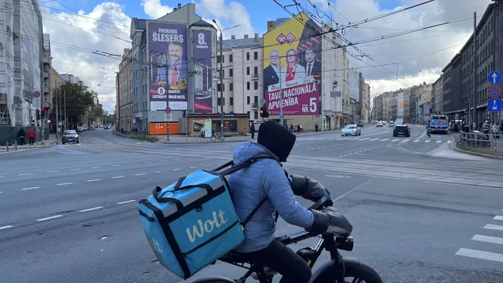 Курьер на электровелосипеде и с контейнером за плечами на перекрестке, на заднем плане видны дома с огромными предвыборными плакатами