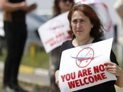 Женщина с плакатом You are not welcome и изображением перечеркнутого самолета в цветах российского флага